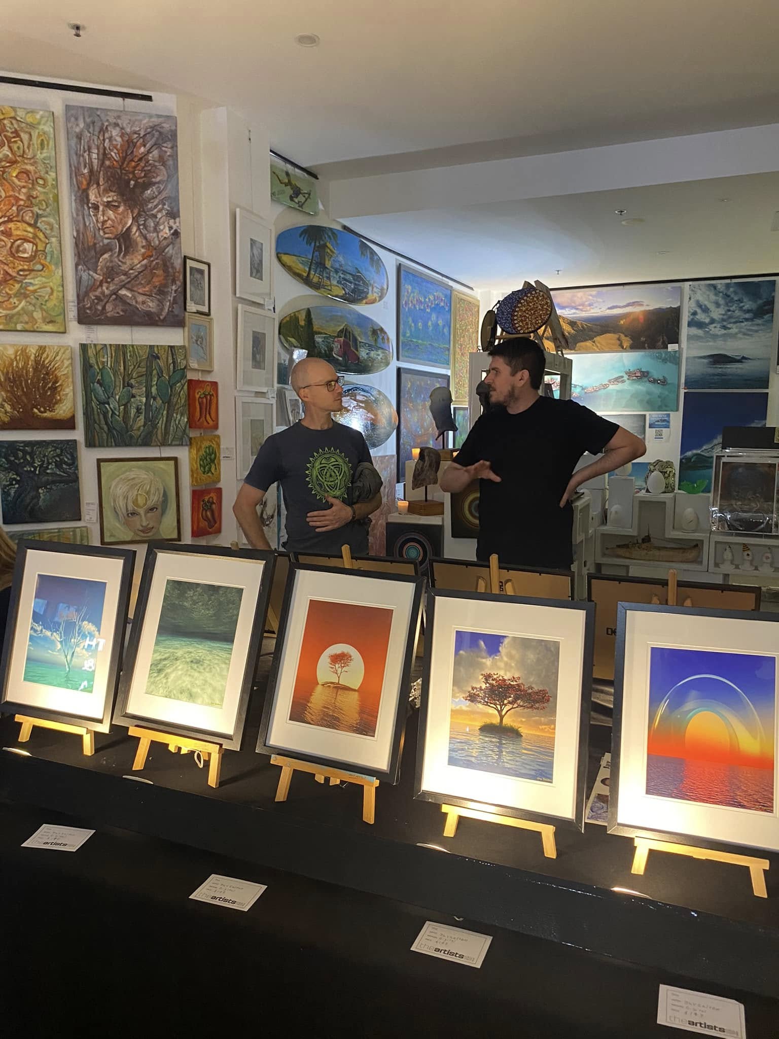 Jay Salton speaking with a visitor beside a display of framed artworks